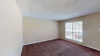 This single-floor residence features two bedrooms and two full bathrooms within a well-organized layout. The living and dining area is carpeted in a deep brown tone, complemented by neutral beige walls and a textured popcorn ceiling. A large window allows natural light to flow into the main living space, providing views of the surrounding exterior. A pendant light fixture above the dining area adds a subtle design touch, while an open passageway connects the living space directly to the kitchen.

The kitchen is equipped with white cabinetry, light-toned countertops, a white electric coil range, and a refrigerator. A pass-through window connects the kitchen to the living and dining area, enhancing the open feel of the space.

Both bedrooms are comfortably sized with the same dark carpet and neutral wall tones found throughout the unit. Each room benefits from window access, providing natural light and outdoor views. The bedrooms are well-suited for furnishing with standard bedroom sets.

The two bathrooms each offer functional layouts. One features a tub-shower combination with chrome fixtures and a light countertop vanity with white cabinetry. The second bathroom mirrors a similar design with tile flooring and a shower-curtain enclosure. Overall, the unit maintains a clean, neutral aesthetic throughout.