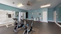 This fitness center is a comprehensively equipped exercise facility spread across a single floor, designed to support a wide range of workout routines. The space is defined by its teal-painted walls and durable rubber flooring, creating a clean, energetic environment conducive to physical activity.

The cardio zone is outfitted with multiple treadmills, elliptical trainers, and a stationary bike, all positioned along walls lined with windows fitted with white blinds. These windows allow ample natural light to fill the space while offering views of the surrounding greenery outdoors. Overhead, ceiling fans and fluorescent lighting ensure the area remains well-lit and comfortable during workouts.

A dedicated strength training area houses a multi-station cable machine by TuffStuff, ideal for a full-body resistance workout. Complementing this is a dumbbell rack offering a range of free weights, along with a set of Stages-branded spin bikes for high-intensity cycling sessions.

The walls are adorned with informational fitness charts and exercise guides, adding a functional and educational element to the space. A water station and additional amenities are conveniently accessible near the entrance. The overall layout is thoughtfully organized, maximizing floor space while providing a diverse array of equipment to accommodate users of all fitness levels.