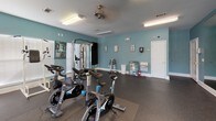 This fitness center is a comprehensively equipped exercise facility spread across a single floor, designed to support a wide range of workout routines. The space is defined by its teal-painted walls and durable rubber flooring, creating a clean, energetic environment conducive to physical activity.

The cardio zone is outfitted with multiple treadmills, elliptical trainers, and a stationary bike, all positioned along walls lined with windows fitted with white blinds. These windows allow ample natural light to fill the space while offering views of the surrounding greenery outdoors. Overhead, ceiling fans and fluorescent lighting ensure the area remains well-lit and comfortable during workouts.

A dedicated strength training area houses a multi-station cable machine by TuffStuff, ideal for a full-body resistance workout. Complementing this is a dumbbell rack offering a range of free weights, along with a set of Stages-branded spin bikes for high-intensity cycling sessions.

The walls are adorned with informational fitness charts and exercise guides, adding a functional and educational element to the space. A water station and additional amenities are conveniently accessible near the entrance. The overall layout is thoughtfully organized, maximizing floor space while providing a diverse array of equipment to accommodate users of all fitness levels.
