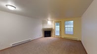 This single-floor space centers around a generously sized living room that immediately draws attention with its clean, open layout and warm, neutral tones. The room is finished with wall-to-wall carpet in a soft beige hue, providing a comfortable and cohesive foundation throughout. A corner fireplace serves as a natural focal point, framed by a simple white mantel that complements the room's understated aesthetic. Natural light filters in through a glass-panel exterior door and an adjacent window, both set against warm yellow-toned walls that add a subtle contrast to the predominantly white interior. Blinds on the window offer privacy and light control as needed. Baseboard heating units run along the lower walls, ensuring efficient and consistent warmth throughout the space. Overhead, a flush-mount light fixture provides direct illumination to the central area of the room. The walls are clean and neutral, offering a versatile canvas for personalization. The overall ambiance is calm and functional, with a practical layout that balances comfort with simplicity — making it a welcoming and easy-to-maintain living environment.