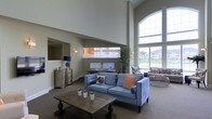 Located at 130 Ocean Aisle Circle in Salisbury, Maryland, this welcoming community clubhouse offers a thoughtfully designed single-level space that blends modern style with comfortable functionality. The interior immediately impresses with its dramatic soaring ceilings, which lend an airy, open atmosphere throughout. Large arched windows flood the space with generous natural light, offering views of the surrounding community and creating a bright, inviting ambiance. The main lounge area is furnished with plush seating, including a contemporary blue sofa accented with decorative pillows, arranged around a rustic wood coffee table — perfect for relaxed gatherings. A dedicated computer workstation area provides residents with convenient access to digital resources. Elegant touches such as wall sconces, a classic dresser with a decorative mirror, and lush indoor plants add warmth and character to the space. A secondary seating nook near the windows features additional casual furniture, ideal for conversation or quiet relaxation. Neutral tones in warm greige and soft gray carry throughout the walls and carpeted floors, creating a cohesive and calming aesthetic. Overall, the space strikes a balance between polished community amenity and comfortable retreat.