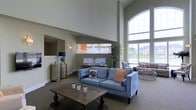 Located at 130 Ocean Aisle Circle in Salisbury, Maryland, this welcoming community clubhouse offers a thoughtfully designed single-level space that blends modern style with comfortable functionality. The interior immediately impresses with its dramatic soaring ceilings, which lend an airy, open atmosphere throughout. Large arched windows flood the space with generous natural light, offering views of the surrounding community and creating a bright, inviting ambiance. The main lounge area is furnished with plush seating, including a contemporary blue sofa accented with decorative pillows, arranged around a rustic wood coffee table — perfect for relaxed gatherings. A dedicated computer workstation area provides residents with convenient access to digital resources. Elegant touches such as wall sconces, a classic dresser with a decorative mirror, and lush indoor plants add warmth and character to the space. A secondary seating nook near the windows features additional casual furniture, ideal for conversation or quiet relaxation. Neutral tones in warm greige and soft gray carry throughout the walls and carpeted floors, creating a cohesive and calming aesthetic. Overall, the space strikes a balance between polished community amenity and comfortable retreat.