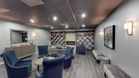 The Quincy Library is a thoughtfully designed single-floor lounge space that blends functionality with refined contemporary style. The room is anchored by a sleek electric fireplace set into a textured accent wall, flanked on both sides by striking diamond-pattern built-in shelving units that display books and decorative items in a visually dynamic arrangement. The shelving design serves as a focal architectural element, adding depth and geometric interest to the space.

The color palette is sophisticated, combining cool grey walls with a darker coffered ceiling that adds dimension overhead. Recessed lighting and wall-mounted sconces provide layered illumination throughout. The flooring consists of warm hardwood-style planks that contrast elegantly with the room's cooler tones.

Seating is generous and well-curated, featuring navy blue upholstered armchairs with nailhead trim arranged around a round side table, alongside a neutral bench and additional accent seating. Decorative touches, including an orchid plant, a framed wall print, and a mirrored side table, contribute to the space's polished, welcoming ambiance. Overall, the Quincy Library offers an intimate yet stylish environment ideal for reading and relaxation.
