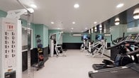 This fitness facility is designed to provide a comprehensive workout environment within a single-level layout. The space features a clean, modern aesthetic highlighted by teal-painted walls with a contrasting black lower panel, creating a visually distinct and energizing atmosphere. Recessed lighting in the white ceiling ensures bright, even illumination throughout.

The room is furnished with a range of strength-training machines, including multi-function cable and resistance equipment suitable for targeting various muscle groups. A dedicated medicine ball rack along one wall offers additional options for functional and dynamic training. Cardio equipment, including a treadmill, is also present, providing users with well-rounded exercise options.

A floor-to-ceiling mirrored wall along one side of the room visually expands the space and allows users to monitor their form during workouts. Mounted flat-screen televisions are positioned for easy viewing, adding an entertainment element to the exercise experience. The light-colored flooring is clean and durable, well-suited to the demands of a fitness environment. Overall, the space is thoughtfully organized to maximize usability while maintaining an open and uncluttered feel.