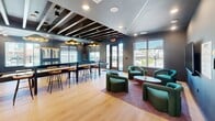 Located at 2418 Main St in Rocky Hill, Connecticut, this contemporary community lounge offers a thoughtfully designed, open-plan social space that blends entertainment, relaxation, and functional amenities seamlessly. The single-floor layout is unified by warm light wood flooring, dark accent walls, and an eye-catching exposed beam ceiling accented with distinctive cloud-shaped pendant lighting and recessed fixtures that create a dynamic, modern ambiance.

The game area is a standout feature, equipped with a full-size pool table and a foosball table, positioned beneath large windows that invite an abundance of natural light and outdoor views. Adjacent to this zone, a long bar-height communal table with wooden stools provides an ideal gathering spot for casual socializing.

A cozy lounge area features plush, teal curved armchairs arranged around a round coffee table on a warm-toned area rug, offering a comfortable retreat. The space also includes a well-appointed kitchenette outfitted with stainless steel appliances, ample cabinetry, a sink, and stylish stone-look tile backsplash — ideal for hosting events.

Dual flat-screen TVs mounted above the kitchenette counter add to the entertainment options. Glass-panel doors provide a seamless transition to adjacent spaces, while large windows throughout ensure the room remains bright and connected to the outdoors.