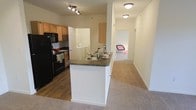 This single-floor residence offers a practical and well-organized layout designed for comfortable everyday living. The kitchen serves as a central hub of the home, featuring dark countertops, light wood cabinetry, and black appliances including a refrigerator, stove, and built-in microwave. A peninsula-style breakfast bar extends from the kitchen, providing additional prep space and a casual dining option while maintaining an open connection to the rest of the living area. Track lighting overhead ensures the kitchen remains bright and functional. Conveniently tucked just off the kitchen is an in-unit laundry area, adding a practical touch to the home's design. The flooring transitions from durable hardwood-style planks in the kitchen to soft, neutral-toned carpet that extends through the living space and into the bedroom, creating a warm and inviting atmosphere. A hallway leads from the main living area to the bedroom, which is visible in the background and benefits from natural light. The overall color palette of soft whites and warm neutrals throughout the unit contributes to an airy, clean aesthetic that feels both welcoming and move-in ready.