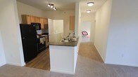 This single-floor residence offers a practical and well-organized layout designed for comfortable everyday living. The kitchen serves as a central hub of the home, featuring dark countertops, light wood cabinetry, and black appliances including a refrigerator, stove, and built-in microwave. A peninsula-style breakfast bar extends from the kitchen, providing additional prep space and a casual dining option while maintaining an open connection to the rest of the living area. Track lighting overhead ensures the kitchen remains bright and functional. Conveniently tucked just off the kitchen is an in-unit laundry area, adding a practical touch to the home's design. The flooring transitions from durable hardwood-style planks in the kitchen to soft, neutral-toned carpet that extends through the living space and into the bedroom, creating a warm and inviting atmosphere. A hallway leads from the main living area to the bedroom, which is visible in the background and benefits from natural light. The overall color palette of soft whites and warm neutrals throughout the unit contributes to an airy, clean aesthetic that feels both welcoming and move-in ready.