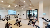 This fitness facility is thoughtfully designed to accommodate a variety of exercise needs across two connected zones. The primary studio is a bright, open space featuring multiple stationary spin bikes arranged for group or individual cycling sessions. Full-length mirrors line the walls, creating an expansive feel while also serving as a functional training aid. A wall-mounted ballet barre runs along the mirrored wall, adding versatility for barre, stretching, or balance-focused workouts. A large flat-screen display is mounted prominently, enabling on-demand or virtual fitness classes. The studio is finished with neutral-toned carpet flooring and features a coffered ceiling with recessed lighting and a decorative pendant ceiling fan, contributing to a clean and inviting atmosphere. Sleek black-framed glass doors connect the studio to a separate weight room, visible through the glazing and filled with strength training equipment. The transparent partition maintains an open, airy quality between the two areas. The overall design is modern and minimalist, with crisp white walls, consistent lighting throughout, and a layout that promotes both functionality and comfort for users at all fitness levels.