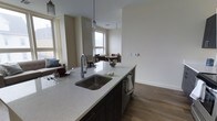 This studio with loft offers a thoughtfully designed open-concept layout that maximizes every inch of its footprint. The kitchen serves as the centerpiece of the main living area, anchored by a large island with a striking quartz countertop featuring subtle speckled detailing, an undermount stainless steel sink, and a sleek pull-down faucet. Dark cabinetry beneath the island provides a bold contrast to the light countertop surface, while pendant lighting above adds a refined, contemporary touch. A dishwasher is seamlessly integrated into the island for added convenience. The open layout flows naturally into the living and dining areas, which benefit from multiple windows that invite generous amounts of natural light, creating a bright and airy atmosphere throughout. Wood-toned flooring runs continuously across the main level, lending warmth and cohesion to the space. The dining area accommodates a small table and chairs, perfectly positioned near the windows. Neutral wall tones throughout keep the aesthetic clean and versatile. The loft component adds a valuable layer of functionality, offering an additional area that can serve as a sleeping space or private retreat above the main living floor.