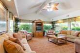Located at 16350 S Harbor Blvd in Santa Ana, California, this single-floor community space offers a welcoming and well-appointed interior designed for comfort and relaxation. The main living area is anchored by soaring vaulted ceilings accented with exposed beams and a decorative ceiling fan, creating an airy, open ambiance throughout. A central structural column subtly defines the generous seating areas, which are furnished with plush sofas and armchairs arranged around a wooden coffee table atop richly patterned carpeting. Large windows dressed with roman shades line multiple walls, flooding the space with natural light and offering pleasant views of the surrounding lush greenery. A built-in wooden entertainment center provides a functional focal point on one side of the room, while framed artwork adds a refined aesthetic touch throughout. Adjacent to the main lounge, a cozy dining area accommodates a round wooden table with upholstered chairs beneath vaulted ceilings, decorated with a pair of elegantly framed botanical prints. Recessed lighting and a ceiling fan ensure comfortable illumination at all times of day. The overall atmosphere is warm, inviting, and thoughtfully curated, making this space well-suited for casual gatherings and everyday comfort.