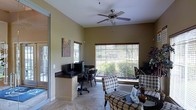 This single-floor space offers a comfortable and functional layout that seamlessly blends a home office with a casual sitting area. The room is filled with natural light streaming through multiple large windows fitted with wooden blinds, offering pleasant views of the surrounding greenery outside. A ceiling fan with a built-in light fixture is centrally mounted, adding both comfort and a subtle decorative touch to the space.

The built-in desk area along one wall provides a dedicated workspace, complete with dark granite-style countertops and ample room for computer equipment and office essentials. A pair of ergonomic office chairs complement the setup, making it a practical area for productivity.

The sitting area is furnished with patterned upholstered chairs arranged around a small side table, creating an inviting nook for relaxation or informal conversation. The neutral, warm-toned walls and light tile flooring tie the space together, giving it a cohesive and airy feel. Decorative artwork adorns the walls, adding a personal touch to the room.

A sliding glass door on one side provides easy access to an outdoor area, further enhancing the connection between the interior and the natural surroundings. Overall, the space strikes a balance between functionality and comfort in a warm, well-lit environment.