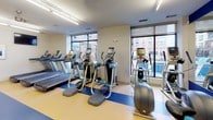 This fitness center is a fully outfitted, single-floor facility designed to accommodate a wide variety of workout routines. The space is visually defined by its striking blue accent walls and matching blue lane striping across light wood-toned flooring, creating a dynamic and energetic atmosphere throughout.

The strength training area is comprehensively equipped with multiple cable machines, including Precor-branded units, a rack of free weights with dumbbells neatly organized along a mirrored wall, Smith machines, adjustable benches, and a variety of resistance stations targeting different muscle groups. Mirrors line portions of the walls, enhancing the sense of space and allowing users to monitor their form.

The cardio zone is positioned near a wall of floor-to-ceiling windows, flooding the area with natural light and offering views of the surrounding outdoor environment, including what appears to be a pool area. This section houses a row of treadmills, elliptical trainers, and step machines, all positioned to take advantage of the natural light and views.

Overhead, bright fluorescent strip lighting ensures the entire floor is evenly illuminated, while ceiling fans contribute to air circulation and comfort. The space also features double-door access, recessed speakers, and a mounted television, rounding out the facility's amenities for a complete fitness experience.