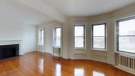 This single-floor space showcases a timeless, classic character defined by rich, polished hardwood floors that stretch throughout the open main living area. The room is bathed in natural light, courtesy of multiple double-hung windows dressed with horizontal blinds, including a distinctive bay window configuration that creates a graceful architectural focal point and offers views of the surrounding neighborhood. The white-painted walls and crisp window trim lend a clean, airy aesthetic that complements the warmth of the gleaming wood floors beneath. A traditional cast-iron radiator is tucked neatly beneath one of the windows, adding a charming, old-world detail consistent with classic urban residential architecture. The open layout provides a flexible and welcoming living environment, with an adjacent doorway visible from the main room, hinting at additional spaces beyond. The combination of abundant windows, well-maintained original flooring, and architectural details such as the bay window and wall moldings give this space a sense of enduring character and comfortable livability, making it an inviting and functional environment throughout all seasons.