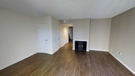 This two-story space offers a straightforward and functional layout with thoughtful design details. The main living area is highlighted by rich, dark wood-style flooring that extends throughout, creating a warm and cohesive look underfoot. A fireplace with a clean white surround serves as a natural focal point in the living space, adding both character and comfort to the room.

Neutral gray walls complement the flooring and contribute to a calm, versatile atmosphere that adapts well to a variety of décor styles. A textured ceiling adds subtle visual interest overhead. Multiple interior doors off the main living area suggest convenient access to closets or additional rooms, while a staircase visible in the background leads to the second floor, indicating a well-organized multi-level floor plan.

Ventilation is well-considered, with ceiling vents positioned throughout the main level. The overall aesthetic is clean and move-in ready, with white trim and door frames providing crisp contrast against the neutral walls. The space strikes a balance between practicality and comfort, making it a versatile option for a range of lifestyles.