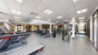 Located at 500 E Constitution Dr in Palatine, IL, this well-appointed fitness center offers a generous, open-plan exercise space spread across a single floor. The facility is designed to accommodate a wide variety of workout routines, with distinct zones dedicated to cardiovascular training, strength equipment, and functional fitness.

The cardio area features treadmills, elliptical machines, a stair climber, and a rowing machine, positioned near floor-to-ceiling windows that flood the space with natural light and provide pleasant outdoor views. The strength training zone is equipped with a full array of weight machines, cable systems, and free weight benches, set against a textured stone-style accent wall that adds visual character to the interior.

A dedicated functional training area includes stability balls, resistance equipment, and a wall-mounted mirror that enhances the sense of openness. The facility also features a mounted flat-screen television for entertainment during workouts. Throughout the space, durable rubber flooring ensures safety and comfort, while a suspended drop-tile ceiling with recessed and panel lighting provides consistent, bright illumination. A glass-paneled entry door connects the fitness center to adjacent common areas, contributing to a cohesive and accessible facility layout.