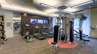 This fitness center is thoughtfully designed to support a full range of workout routines in a clean, modern environment. The space features a row of treadmills positioned in front of wall-mounted flat-screen televisions, allowing users to stay entertained during cardio sessions. An elliptical trainer is also available, offering a low-impact alternative for cardiovascular exercise. On the strength training side, a multi-station cable machine — branded Magnum — provides versatile resistance options for a comprehensive full-body workout. The flooring combines patterned carpet with a bold circular accent area in warm tones beneath the strength equipment, adding visual interest and defining workout zones. Overhead, a layered tray ceiling with recessed linear lighting creates a bright, polished atmosphere, while mirrored wall panels expand the sense of space and allow users to monitor their form. The warm wood-paneled accent wall frames the television display area, lending the room a refined, upscale aesthetic. Natural light filters in through a window, complementing the well-designed interior lighting throughout this inviting and functional fitness facility.