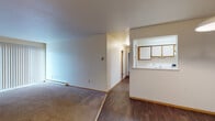 This single-floor unit offers a practical and functional layout designed for comfortable everyday living. The living area is generously sized, featuring soft carpet flooring, a large sliding glass door dressed with vertical blinds that allows natural light to fill the space, and a baseboard heating system along the perimeter. The transition from carpet to dark wood-style hard flooring marks the shift into the dining and kitchen areas.

The kitchen is accessible via a pass-through counter opening, providing a connected feel between the cooking and living spaces. It is equipped with wood-toned upper cabinetry, a coil-burner electric range with overhead ventilation, a sink, and a refrigerator, all finished in white.

A hallway connects the main living area to the private quarters. The bedrooms feature plush carpet flooring, neutral walls, individual windows with blinds for natural light, baseboard heaters, and wall-mounted air conditioning units for year-round climate control. Closet storage is also present.

The bathroom is well-appointed with a full-size soaking tub and tiled surround, a single vanity with countertop sink, wood-toned cabinetry, and dark wood-style flooring that ties in with the rest of the unit. The entry foyer features a six-panel door with warm wood trim framing, with an adjacent closet providing convenient storage upon arrival.