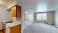 This single-floor unit offers a practical and straightforward layout designed for comfortable everyday living. The kitchen is efficiently arranged in a galley-style configuration, featuring warm oak cabinetry, a white electric coil range, refrigerator, sink, and under-cabinet range hood. Light wood-look flooring runs throughout the kitchen, providing a clean, easy-to-maintain surface.

The living area is open and well-proportioned, finished with neutral gray carpet and light gray walls that create a calm, understated atmosphere. A large window along the exterior wall allows natural light to fill the space while offering outdoor views, and a wall-mounted air conditioning unit provides climate control. The layout connects the living area directly to the kitchen, supporting an open flow between the two spaces.

The bedroom is a comfortable, quiet room also finished with plush carpet and neutral tones. A window provides additional natural light, and a closet offers convenient built-in storage. The full bathroom features a tiled tub and shower combination, toilet, and vanity sink, finished with wood-look flooring that complements the rest of the unit. Overall, the space presents a clean, move-in ready environment with a cohesive, neutral aesthetic throughout.