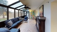 This single-floor space offers a thoughtfully designed layout that blends comfort and functionality across several distinct areas. The standout feature is a spacious sunroom enclosed by floor-to-ceiling glass panels and a sloped glass skylight roof, allowing abundant natural light to flood the interior throughout the day. The sunroom is finished with warm brick-patterned tile flooring and is equipped with a ceiling fan, making it a comfortable retreat in any season. A decorative fireplace mantle and a colorful folding room divider add distinctive character to the space, while blue cushioned patio furniture provides inviting seating. An adjacent screened enclosure extends the outdoor-living experience, offering wide views of surrounding greenery and mature trees. The interior common area features patterned carpet flooring, a drop ceiling with recessed lighting, and a connected kitchenette complete with cabinetry, countertops, and a dining area with bar seating. Upholstered lounge chairs are arranged to encourage conversation and relaxation. Additional utility areas include textured stucco walls, security camera installations, and practical access panels. Together, these spaces create a well-rounded environment suited for both leisure and everyday activity.