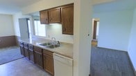 This single-story space presents a practical and straightforward layout, with the kitchen serving as a central functional hub. The kitchen features rich, dark wood cabinetry both above and below the counter, providing ample storage. A double stainless steel sink is positioned beneath a window, allowing natural light to brighten the workspace. Light-toned laminate countertops run along the cabinetry, and a white dishwasher is integrated beneath the counter for added convenience. The tile flooring in the kitchen offers durability and easy maintenance.

A half-wall partition separates the kitchen from the adjacent living area, which transitions to carpeted flooring and is finished in neutral, light tones. This open sightline between the two areas creates a connected, airy feel throughout the main living space. A doorway visible from the kitchen leads further into the unit, suggesting additional rooms beyond. The overall color palette throughout is soft and neutral, with white walls and warm wood accents contributing to a clean, understated ambiance. The space is well-suited for straightforward, everyday living.