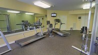 This fitness center offers a practical and functional space for cardiovascular exercise and general wellness. The room is outfitted with multiple treadmills and a stationary upright bike, providing a solid selection of cardio equipment to accommodate various workout preferences. A large wall-mounted mirror spans one side of the room, visually expanding the space and allowing users to monitor their form during exercise. A flat-screen television is mounted on the wall, offering entertainment to keep workouts engaging. The flooring consists of durable rubber-style gym matting, providing cushioning and stability underfoot. Overhead, flush-mounted lighting ensures the room is well-lit throughout. The walls are painted in a soft yellow-green tone, contributing to a light and energizing atmosphere. A door at the rear of the room provides additional access or ventilation. The overall layout is open and uncluttered, allowing for comfortable movement between equipment. This single-level fitness facility is thoughtfully designed to serve the health and wellness needs of its users in a clean, organized, and welcoming environment.