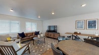 This single-floor communal space is thoughtfully designed to accommodate a variety of social and functional needs. The open layout is divided into distinct zones that flow seamlessly into one another, creating a welcoming and versatile environment.

The lounge area serves as the centerpiece of the space, furnished with a rich brown leather sofa, accent chairs, and a patterned area rug that anchors the seating arrangement. A large flat-screen television mounted on a rustic wood media console provides a focal point, while recessed lighting and a window with blinds contribute to a comfortable, well-lit atmosphere.

Adjacent to the lounge, a dedicated dining and gathering zone features both a high-top communal table with bar stools and a round dining table with upholstered chairs, offering flexible seating options for groups of various sizes. Warm hardwood-style flooring runs throughout this area, complemented by neutral wall tones and framed artwork that add a polished, inviting touch.

The kitchenette is equipped with dark wood cabinetry, light countertops, a stainless steel refrigerator, a microwave, a sink, and a coffee maker—providing essential amenities for events or everyday use. A separate entry door and multiple windows allow natural light to fill the space while maintaining easy access and circulation throughout.
