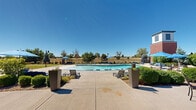 Located in Denver, CO, this outdoor pool area is a well-appointed amenity space designed for relaxation and recreation. At its centerpiece is a generously sized freeform swimming pool with a smooth blue-tiled edge and built-in entry steps, allowing for easy access from multiple points. The surrounding concrete sundeck provides ample room for lounging, with a selection of sling-style chaise lounge chairs positioned throughout to take full advantage of Colorado's abundant sunshine and open blue skies.

Adjacent to the pool, a dedicated patio area features round tables with chairs, offering a comfortable setting for outdoor dining or casual gathering. Shade is provided by bold blue market umbrellas stationed across the deck, creating inviting spots to relax out of direct sunlight. Neatly trimmed hedges, ornamental planters with seasonal flowers, and mature trees frame the space, adding a layer of greenery that softens the hardscape surroundings.

The pool area is bordered by a low perimeter fence that opens to views of the open Colorado landscape and treeline beyond. A distinctive brick and white-paneled tower structure anchors one corner of the space, complementing the architecture of the adjacent multi-story building, which features a mix of brick, white siding, and large glass windows. A covered entry with brick detailing and a teal door provides access to the connected facility, completing this cohesive and functional outdoor amenity space.