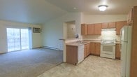 This single-floor unit features an open-concept layout that connects the kitchen and living area, creating a cohesive and functional living space. The kitchen is equipped with medium-toned wood cabinetry, light laminate countertops, a peninsula with an integrated sink and dishwasher, and a white electric range with an overhead exhaust hood. The peninsula serves as a natural divider between the kitchen and the adjoining living space, offering additional prep or casual seating space. The kitchen flooring is a neutral vinyl tile, providing durability and ease of maintenance. The living area transitions to wall-to-wall carpeting, adding warmth and comfort underfoot. A baseboard heating unit runs along the wall, while a wall-mounted air conditioning unit provides climate control. The vaulted or angled ceiling detail in the living room adds a subtle architectural element, creating a slightly more open feel overhead. Soft, neutral wall tones throughout contribute to a clean and versatile aesthetic. A doorway visible beyond the living area suggests additional rooms or storage space within the unit.