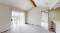 This single-story residence offers a thoughtfully arranged layout with distinct living, sleeping, and functional spaces. The living area is a standout, featuring vaulted ceilings accented by an exposed wood beam that adds architectural character. A full-width sliding glass door fitted with vertical blinds fills the space with natural light and provides views of the surrounding exterior. Soft carpeting throughout the living and bedroom areas contributes to a comfortable, quiet atmosphere.

Adjacent to the living area, a pass-through kitchen opens via a countertop window, creating an open, connected feel. The kitchen is equipped with black appliances including a range, over-the-range microwave, and refrigerator, complemented by white cabinetry and a geometric tile backsplash. Light wood-style flooring carries through the kitchen and hallway.

Built-in open shelving beside the kitchen pass-through provides convenient display or storage space. A hallway connects the living areas to additional rooms, including a bedroom outfitted with a ceiling fan and carpeted flooring. The bathroom features a full-sized tub-shower combination with a clean white surround and wood-style flooring. The textured ceilings are consistent throughout the unit.