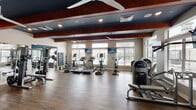Located at 4500 E Ray Rd in Gilbert, AZ, this well-appointed fitness center offers a comprehensive workout environment designed to accommodate a wide range of exercise needs. The single-floor facility is organized into distinct zones, each tailored to specific training styles.

The main cardio area features a row of treadmills and elliptical machines positioned along a wall of large windows fitted with blinds, allowing abundant natural light to fill the space while providing views of the surrounding landscape. Wall-mounted televisions are positioned throughout for entertainment during workouts. The ceilings are finished in a deep navy-blue tone and accented by rich wood beams, lending the space a modern yet warm aesthetic. Ceiling fans throughout ensure comfortable airflow.

A dedicated strength training zone houses a multi-station cable machine and functional trainer setup, surrounded by mirrored walls and durable wood-look flooring. This area offers ample open floor space for stretching and dynamic movement exercises.

The free weights area features a textured, dark accent wall paired with full-length mirrors and organized dumbbell racks ranging across multiple weight levels, complemented by an adjustable workout bench. A separate barbell and plate storage section rounds out the weight training offerings.

Overall, the fitness center combines practical functionality with polished interior design, creating a motivating and well-equipped environment for residents and users in Gilbert, AZ.