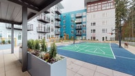 This outdoor amenity space is thoughtfully designed to serve as a central gathering hub within a modern multi-story residential community. The courtyard is anchored by a teal-surfaced sport court with clearly marked court lines, set against a backdrop of contemporary buildings clad in teal, gray, and white panels with private balconies.

A covered pavilion provides a sheltered focal point, featuring a dark-paneled ceiling, sturdy structural columns, and slatted wood privacy screens that filter light while defining the space. Beneath the canopy sits a striking blue outdoor kitchen island equipped with a sink and ample prep space, complemented by comfortable lounge seating arranged for relaxed conversation.

Adjacent to the pavilion, a spacious open-air patio hosts multiple dining tables and chairs illuminated by strung bistro lights overhead, creating an inviting evening atmosphere. Raised planters with ornamental grasses, shrubs, and decorative globe accents add greenery and visual interest throughout. Smooth large-format concrete pavers unify the entire outdoor area, while mature trees visible beyond the perimeter provide a natural backdrop. The overall ambiance is modern, active, and community-oriented.