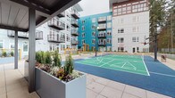 This outdoor amenity space is thoughtfully designed to serve as a central gathering hub within a modern multi-story residential community. The courtyard is anchored by a teal-surfaced sport court with clearly marked court lines, set against a backdrop of contemporary buildings clad in teal, gray, and white panels with private balconies.

A covered pavilion provides a sheltered focal point, featuring a dark-paneled ceiling, sturdy structural columns, and slatted wood privacy screens that filter light while defining the space. Beneath the canopy sits a striking blue outdoor kitchen island equipped with a sink and ample prep space, complemented by comfortable lounge seating arranged for relaxed conversation.

Adjacent to the pavilion, a spacious open-air patio hosts multiple dining tables and chairs illuminated by strung bistro lights overhead, creating an inviting evening atmosphere. Raised planters with ornamental grasses, shrubs, and decorative globe accents add greenery and visual interest throughout. Smooth large-format concrete pavers unify the entire outdoor area, while mature trees visible beyond the perimeter provide a natural backdrop. The overall ambiance is modern, active, and community-oriented.