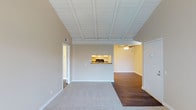 This single-floor residence offers a thoughtfully designed layout that balances openness with defined living areas. The main living space immediately draws attention with its striking vaulted ceiling adorned with white-painted exposed beams, giving the room a sense of elevated volume and architectural character. Soft, neutral wall tones in warm beige create a calm, welcoming ambiance throughout.

The living area features plush wall-to-wall carpeting that transitions smoothly into rich hardwood flooring in the adjacent room, offering both comfort and visual contrast. A pass-through window connects the living space to a secondary area, adding a functional and charming architectural detail that also allows light to flow between rooms.

The adjoining room, illuminated by a ceiling fan with integrated lighting, provides additional living or dining space and continues the warm, neutral color palette. The open sightlines between areas enhance the overall sense of spaciousness, making the layout feel expansive and connected.

Additional features include visible HVAC vents integrated cleanly into the walls, white baseboards and door trim that provide crisp contrast to the neutral walls, and multiple doorways that suggest well-connected interior circulation. The overall design aesthetic is clean, understated, and move-in ready, making it a versatile and functional space.