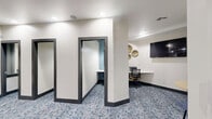 Located at 10107 Ancora Cir in Orlando, FL, this well-appointed business center offers a thoughtfully designed workspace suited for both focused productivity and relaxed collaboration. The space is organized across a single floor and comprises three private office pods alongside a welcoming lounge area.

The private offices are accessed through dark-framed glass panel doors, each pod equipped with a built-in desk and task chair. One pod features teal accent walls and pendant lighting, lending a distinct, focused atmosphere. Interior windows with dark trim frames connect the office pods visually to the lounge, promoting an open yet structured feel throughout.

The lounge area serves as a central gathering point, furnished with warm cognac leather armchairs arranged around a round dark-topped coffee table, accented by potted greenery. A wall-mounted flat-screen TV and decorative gold wall art add functional and aesthetic appeal to this seating zone. A high-top table with bar stools and patterned curtains flanking a sliding glass door provide access to an outdoor courtyard, bringing in natural light and views of the exterior amenity area.

Throughout the space, a patterned blue-and-white carpet adds a modern, professional touch, complementing the crisp white walls and recessed ceiling lighting. The overall ambiance is clean, contemporary, and purposefully designed to support a variety of work styles.