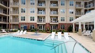 Located at 4800 Marriott Dr in Raleigh, NC, this outdoor amenity space is designed to deliver a resort-like experience within a multi-story residential community. The architecture features a classic combination of brick and neutral-toned siding, with multiple balconied floors overlooking the well-appointed grounds below.

The centerpiece of the outdoor area is a generously sized swimming pool with crystal-clear blue water, framed by a wide pool deck lined with modern white chaise lounges. A decorative water feature with a fountain adds an elegant focal point near the pool's edge, complemented by manicured landscaping, potted topiaries, and flowering plants that enhance the overall aesthetic.

Adjacent to the pool, a spacious paved courtyard serves as an al fresco dining and gathering space. Multiple round tables paired with sleek aluminum chairs are shaded by bold blue-and-white striped market umbrellas, creating a vibrant and inviting atmosphere. A built-in brick grilling station outfitted with a stainless steel grill and granite countertop provides a dedicated area for outdoor cooking and entertaining.

A separate lounge zone features contemporary dark-framed sofas with plush white cushions, positioned alongside decorative stone planters and subtle ground-level lighting, offering a more relaxed and intimate setting. Large windows and glass doors along the building's ground-floor facade connect the interior spaces to the outdoor environment, reinforcing a seamless indoor-outdoor living experience.