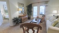 This single-floor space exudes a comfortable, classic charm through its thoughtfully curated design and warm, inviting ambiance. The living area serves as the heart of the space, anchored by a glass-topped coffee table and complemented by a natural wicker accent chair that adds a relaxed, organic feel. A richly toned wooden dresser topped with an elegant table lamp and ornate candlesticks sits beneath a decorative silver-framed mirror, lending a refined, traditional character to the room. Lush indoor plants, including a tall potted palm and a full leafy floor plant, bring vibrant greenery indoors and create a refreshing, nature-inspired atmosphere. French doors with multi-pane glass panels flood the space with abundant natural light and offer views of the surrounding outdoor greenery, creating a seamless connection between the interior and the outdoors. Soft, neutral-toned carpeting throughout the living area adds warmth and comfort underfoot. The overall aesthetic blends traditional furnishings with natural textures and botanical accents, resulting in a space that feels both welcoming and timeless.