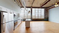 This single-floor industrial loft showcases a compelling mix of historic architectural details and contemporary finishes. The expansive main living area is defined by original hardwood floors, soaring wood-plank ceilings supported by weathered timber beams, and dramatic exposed brick walls bearing layers of aged paint that speak to the building's rich history. A large bank of industrial-style multi-pane windows floods the space with natural light and offers generous outdoor views, enhancing the open, airy atmosphere.

The kitchen is thoughtfully appointed with white shaker cabinetry, granite countertops, stainless steel appliances including a refrigerator, range, microwave, and dishwasher, and a central island. Pendant lighting and a prominent exposed ductwork run overhead, complementing the industrial aesthetic.

A secondary area features polished concrete floors and raw, heavily textured brick walls with remnants of vibrant blue paint, adding to the space's character. This area connects to an additional room with concrete floors and exposed brick, offering versatile utility. The bathroom is cleanly finished with a white vanity, granite countertop, tile-surround walk-in shower, and hardwood flooring, with an exposed brick accent wall providing a seamless tie to the overall industrial design language throughout.