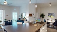 Located in Tampa, Florida, this single-story residence offers a thoughtfully designed open-concept layout with modern finishes throughout. The kitchen is a standout feature, boasting a large island with dark countertops and bar seating, sleek white cabinetry, stainless steel appliances, and pendant lighting that adds a polished, contemporary feel. The adjoining living area is bright and welcoming, with large windows and a glass door that flood the space with natural light while offering views of the surrounding exterior. Light wood-style flooring flows seamlessly throughout the home. A dedicated dining area provides an intimate setting with ample space for a full table and seating. The bedrooms are well-appointed, each offering generous natural light and neutral tones. The primary bathroom features a frameless glass walk-in shower with large-format tile, LED-lit mirrors, and modern grey cabinetry with a clean white countertop. A second bathroom offers similar contemporary styling with a dark-topped vanity and illuminated mirror. The overall ambiance is clean, bright, and modern, with a cohesive design language that balances comfort and style throughout every room.