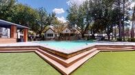 The Tides clubhouse and amenity facility is a single-story community space designed to blend indoor comfort with outdoor leisure. At the heart of the property is a generously sized resort-style pool, elevated on a tiered platform framed by warm wood decking and stone-tiled coping. Manicured turf surrounds the pool area, and a backdrop of mature shade trees provides a lush, park-like atmosphere under open skies. The charming poolhouse structure features a distinctive pitched roof with terracotta tiles, white stucco exterior, and stone accent walls, giving it a refined yet approachable architectural character. Entry to the interior is made through full-length glass French doors framed in slate gray, allowing natural light to flow freely into the space. Inside, light-toned tile flooring carries throughout, and the interior is furnished for recreation and socializing, including a pool table visible from the entrance. Horizontal cedar wood cladding accents the exterior walls near the pool gate, adding a warm, natural texture that complements the surrounding landscape. Iron-railed gated access leads directly to the pool area, reinforcing both safety and style. The overall ambiance is welcoming and vibrant, offering a well-appointed retreat that balances casual recreation with polished design.