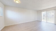 This single-floor unit offers a clean, modern aesthetic throughout, characterized by crisp white walls, light hardwood-style flooring, and textured ceilings that lend a classic residential feel. The main living area is open and airy, bathed in natural light streaming through a sliding glass door that provides access to an outdoor space, creating a seamless connection between interior and exterior. Flush-mounted ceiling fixtures provide consistent overhead lighting throughout.

The galley-style kitchen is efficiently designed with white cabinetry, light countertops, and wood-style flooring. It is equipped with black appliances including a refrigerator and electric range, along with a stainless steel microwave, offering a functional cooking environment.

The bathroom is well-appointed with a combination tub and shower, a white vanity with an integrated sink, and a mirrored medicine cabinet above. Bright vanity lighting enhances the clean, white finish of the space.

Storage is a notable feature, with walk-in closet space offering built-in shelving and hanging rods, carpeted underfoot for comfort. A secondary closet provides additional organizational capacity. Overall, the unit presents a bright, neutral palette that creates a versatile and welcoming living environment.