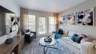 This single-story residence offers a thoughtfully designed layout with modern finishes and a cohesive neutral color palette. The kitchen is outfitted with stainless steel appliances, including a refrigerator and electric range, white shaker-style cabinetry, dark countertops, and wood-look tile flooring that extends a clean, contemporary aesthetic throughout the space.

The living area is generously sized, featuring large windows and glass patio doors that invite abundant natural light and provide outdoor views. The open layout accommodates both a seating arrangement and media space, creating a versatile and comfortable environment.

Two well-proportioned bedrooms each feature large windows with views of surrounding greenery, neutral walls, and ample room for full furnishings. The flooring varies between carpet and hard-surface options, contributing to each room's distinct character.

Two bathrooms mirror each other in design, each featuring white cabinetry, dark countertops, wood-look tile flooring, and a tub-shower combination. The clean, minimal styling ensures a functional and polished finish. Overall, the residence presents a consistent modern aesthetic with practical amenities throughout.