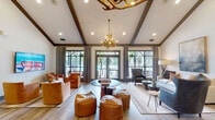 Located at 4095 Lawson Ridge Dr in Huntsville, Alabama, this single-story community clubhouse showcases a thoughtfully designed interior that blends rustic charm with modern comfort. The centerpiece of the space is a grand open lounge area distinguished by soaring vaulted ceilings accented with dark wood beams and dramatic antler chandeliers that serve as striking focal points. Light wood-look flooring flows throughout, complementing the warm, neutral color palette.

The lounge is generously furnished with a mix of leather armchairs, ottomans, a plush sofa, and accent tables, creating multiple inviting seating arrangements suited for socializing or relaxing. Large windows and glass French doors flood the interior with natural light while offering views of the outdoor pool area, seamlessly connecting the indoors to the exterior amenities.

A dedicated kitchen area supports gatherings and events, outfitted with stainless steel appliances including a double wall oven, dishwasher, and refrigerator, set against a distinctive multicolored wood-plank accent wall and dark countertops. A separate coffee station adds a convenient amenity for daily use.

Additional rooms branch off the main lounge, including what appears to be a private office or conference space and a dining area adorned with curated artwork. Throughout the space, carefully selected décor, indoor greenery, and layered lighting contribute to a warm and welcoming ambiance.