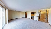 This single-floor residence offers a straightforward and functional layout designed for comfortable everyday living. The spacious main living area is finished with soft, plush wall-to-wall carpeting and neutral-toned walls, creating a clean and versatile space that can be arranged to suit a variety of needs. Standard white ceilings provide a bright overhead finish, while the open feel of the room allows for generous furniture placement and easy movement throughout.

Toward the rear of the living area, an open passthrough leads directly into the kitchen, which features dark espresso-toned cabinetry paired with light countertops, offering a classic contrast. A white range and built-in ventilation hood anchor the cooking space, while the kitchen's efficient layout makes the most of the available area. A raised breakfast bar serves as a natural divider between the kitchen and the adjoining spaces, offering casual seating and added counter space.

Additional doorways visible from the main living area suggest access to further rooms, including what appears to be a hallway leading to additional living spaces. The overall ambiance of the home is practical and understated, with a neutral color palette throughout that lends itself well to personalization and décor.