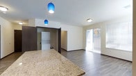 This single-floor unit offers a thoughtfully designed open-concept layout that seamlessly connects the kitchen and living areas. The kitchen is anchored by a generous granite-topped island or breakfast bar, showcasing warm, speckled tones that add a touch of elegance to the space. Two striking cobalt blue pendant lights hang above the counter, providing both functional illumination and a bold design accent that draws the eye.

The open living area features durable wood-look flooring in a rich, dark tone that runs throughout, complemented by clean white walls and dark-framed doorways that create a modern contrast. Natural light flows into the living space through a large sliding glass door fitted with sheer curtains, which opens to an outdoor area, enhancing the sense of openness and connectivity to the exterior.

A dedicated laundry nook is conveniently located just off the main living space, housing a stacked washer and dryer unit — a practical in-unit amenity. The overall layout is clean and efficient, with smooth transitions between functional zones. The neutral palette, modern fixtures, and well-placed lighting combine to create a bright, contemporary ambiance that is both welcoming and practical.