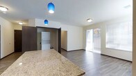 This single-floor unit offers a thoughtfully designed open-concept layout that seamlessly connects the kitchen and living areas. The kitchen is anchored by a generous granite-topped island or breakfast bar, showcasing warm, speckled tones that add a touch of elegance to the space. Two striking cobalt blue pendant lights hang above the counter, providing both functional illumination and a bold design accent that draws the eye.

The open living area features durable wood-look flooring in a rich, dark tone that runs throughout, complemented by clean white walls and dark-framed doorways that create a modern contrast. Natural light flows into the living space through a large sliding glass door fitted with sheer curtains, which opens to an outdoor area, enhancing the sense of openness and connectivity to the exterior.

A dedicated laundry nook is conveniently located just off the main living space, housing a stacked washer and dryer unit — a practical in-unit amenity. The overall layout is clean and efficient, with smooth transitions between functional zones. The neutral palette, modern fixtures, and well-placed lighting combine to create a bright, contemporary ambiance that is both welcoming and practical.