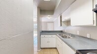 This single-floor unit offers a practical and efficient layout across a modest footprint. The open living and dining area is finished with warm-toned ceramic tile flooring, crown molding, and a ceiling fan with integrated lighting, lending a clean and functional aesthetic. Neutral gray walls and a textured ceiling complement the bright, airy feel, with vertical blinds covering a sliding door or large window that allows natural light to filter in.

The kitchen is thoughtfully arranged in an L-shape configuration, featuring white cabinetry, gray laminate countertops, a stainless steel double sink, and black appliances including a coil-burner electric range with overhead exhaust hood and a full-size refrigerator. A pass-through counter connects the kitchen to the dining and living area, creating an open flow between spaces.

Two bathrooms serve the unit, with at least one featuring tan tile flooring, a tiled tub-surround with shower, toilet, and an overhead storage cabinet. A dedicated closet provides additional storage. A hallway with tile flooring connects the main living areas to the bathrooms and utility spaces, where parquet wood flooring and an electrical panel are also visible. Overall, the unit is designed for straightforward, comfortable living with durable, easy-to-maintain finishes throughout.