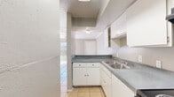 This single-floor unit offers a practical and efficient layout across a modest footprint. The open living and dining area is finished with warm-toned ceramic tile flooring, crown molding, and a ceiling fan with integrated lighting, lending a clean and functional aesthetic. Neutral gray walls and a textured ceiling complement the bright, airy feel, with vertical blinds covering a sliding door or large window that allows natural light to filter in.

The kitchen is thoughtfully arranged in an L-shape configuration, featuring white cabinetry, gray laminate countertops, a stainless steel double sink, and black appliances including a coil-burner electric range with overhead exhaust hood and a full-size refrigerator. A pass-through counter connects the kitchen to the dining and living area, creating an open flow between spaces.

Two bathrooms serve the unit, with at least one featuring tan tile flooring, a tiled tub-surround with shower, toilet, and an overhead storage cabinet. A dedicated closet provides additional storage. A hallway with tile flooring connects the main living areas to the bathrooms and utility spaces, where parquet wood flooring and an electrical panel are also visible. Overall, the unit is designed for straightforward, comfortable living with durable, easy-to-maintain finishes throughout.