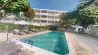 This ground-floor unit is situated within a multi-story building that presents a clean, classic exterior finished in white, with covered balconies lining each level. The building's design reflects a tropical resort-style aesthetic, characterized by structured symmetry and open corridor layouts that promote airflow and natural light throughout the property.

The standout feature of the shared outdoor space is a generously sized rectangular swimming pool with clear turquoise water, framed by a smooth pool deck. A row of cushioned lounge chairs and sun umbrellas lines the poolside, providing dedicated space for relaxation. The surrounding area is complemented by mature tropical trees and manicured hedging, creating a lush, shaded environment that enhances the outdoor living experience.

A decorative iron fence encloses the pool area, adding both security and visual structure to the grounds. Sandy textured surfaces near the perimeter suggest proximity to a coastal or beach-adjacent environment, reinforcing the tropical character of the property.

Interior images were not sufficiently lit to provide detailed descriptions of the unit's indoor spaces. Overall, the property offers a well-maintained, resort-inspired setting suited for those seeking a tropical lifestyle with shared amenity access.