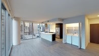 This contemporary high-rise residence offers a thoughtfully designed single-floor open-concept layout that seamlessly blends living, dining, and kitchen spaces. Expansive floor-to-ceiling windows flood the interior with natural light while framing dramatic panoramic views of the surrounding urban skyline — a defining feature of the space.

The kitchen is sleek and modern, anchored by a clean white peninsula with ample counter space ideal for both meal preparation and casual dining. Dark-toned cabinetry provides a striking contrast against the bright, minimalist aesthetic, while stainless steel appliances, including a full-size refrigerator, add a polished finish. A dedicated laundry area is conveniently located just off the kitchen, housing a full-size washer and dryer alongside open shelving for additional storage.

Throughout the space, light-toned hardwood-style flooring enhances the sense of openness and complements the neutral color palette of white and soft gray tones. The smooth, high ceilings further elevate the airy, spacious feel of the interior. The overall ambiance is clean, contemporary, and bright — ideal for those who appreciate modern urban living with sweeping city vistas as an ever-present backdrop.
