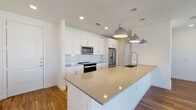 This single-floor unit showcases a thoughtfully designed open-concept layout that seamlessly connects the kitchen and living areas. The kitchen is a standout feature, equipped with crisp white shaker cabinetry, light quartz countertops, and a full suite of stainless steel appliances, including a range, built-in microwave, refrigerator, and dishwasher. A large central island with an undermount sink and industrial-style pendant lighting serves as both a functional workspace and a natural gathering point. A marble-look tile backsplash adds a refined touch throughout the cooking area.

The open living space features warm hardwood-style flooring and recessed lighting, creating a bright and inviting atmosphere. French doors open to an outdoor balcony, allowing abundant natural light to flow in while providing outdoor views. The bedroom is carpeted for comfort and includes multiple windows fitted with blinds, a ceiling fan, and ample natural light.

The bathroom is finished with a white vanity topped with a gray quartz countertop, an illuminated LED mirror, wood-look flooring, and a full tub-shower combo surrounded by neutral tile. Clean lines and a consistent neutral color palette throughout the unit create a cohesive, modern aesthetic.